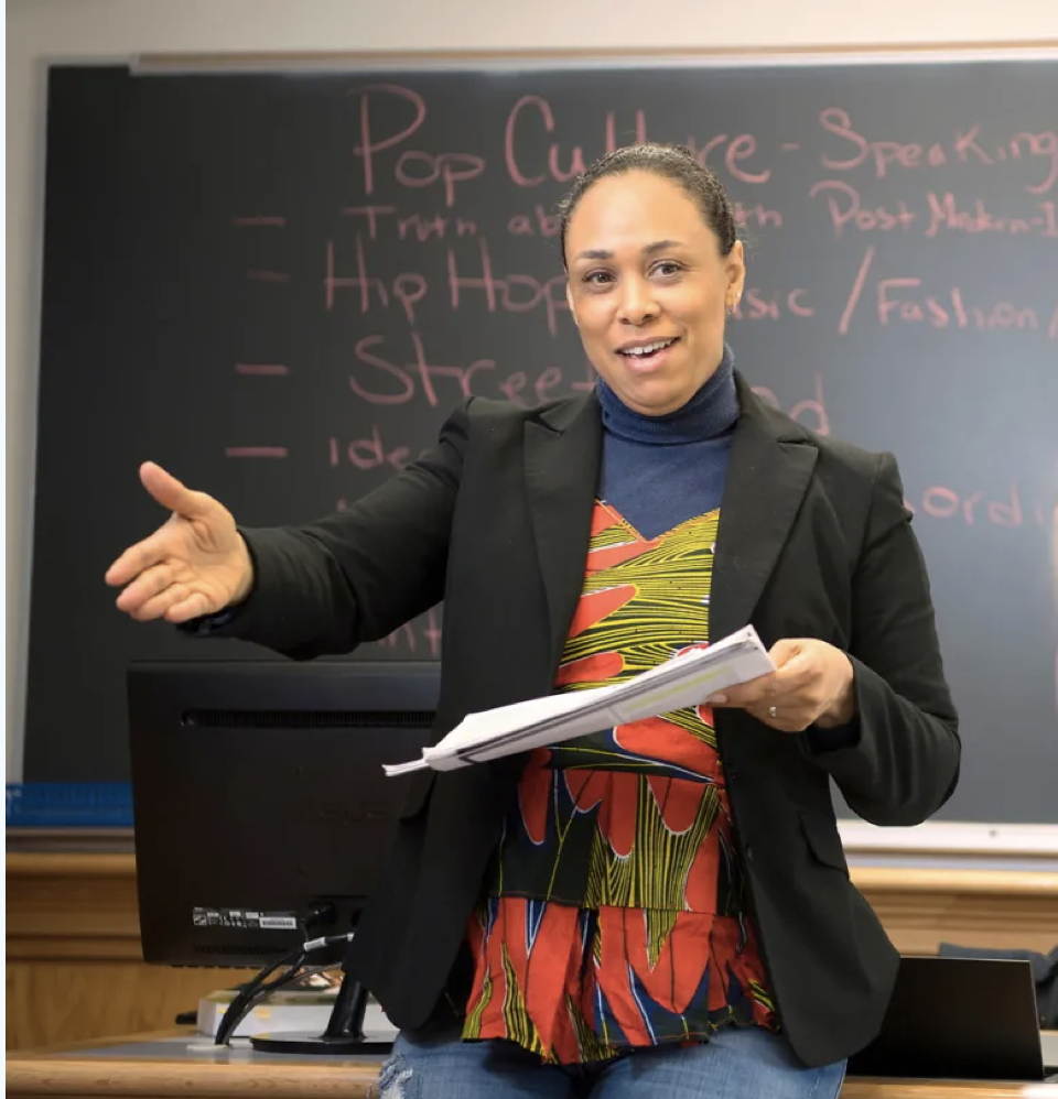 Professor Fourshey, hand extended and speaking to students in the classroom, teaching in front of the Chalkboard with information for her popular Culture in Africa Course written in pink chalk. Her dark brown hair is pulled back in a bun. She is wearing a black blazer over an orange, yellow, and Blue tailored top made in The Gambia.