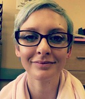 Lisa Wiebesiek sitting in front of her office file cabinet. She is wearing red thick rimmed rectangular framed glasses classes and has a pink scarf around her neck. Her light brown hair is worn in a short close cut pixie style.